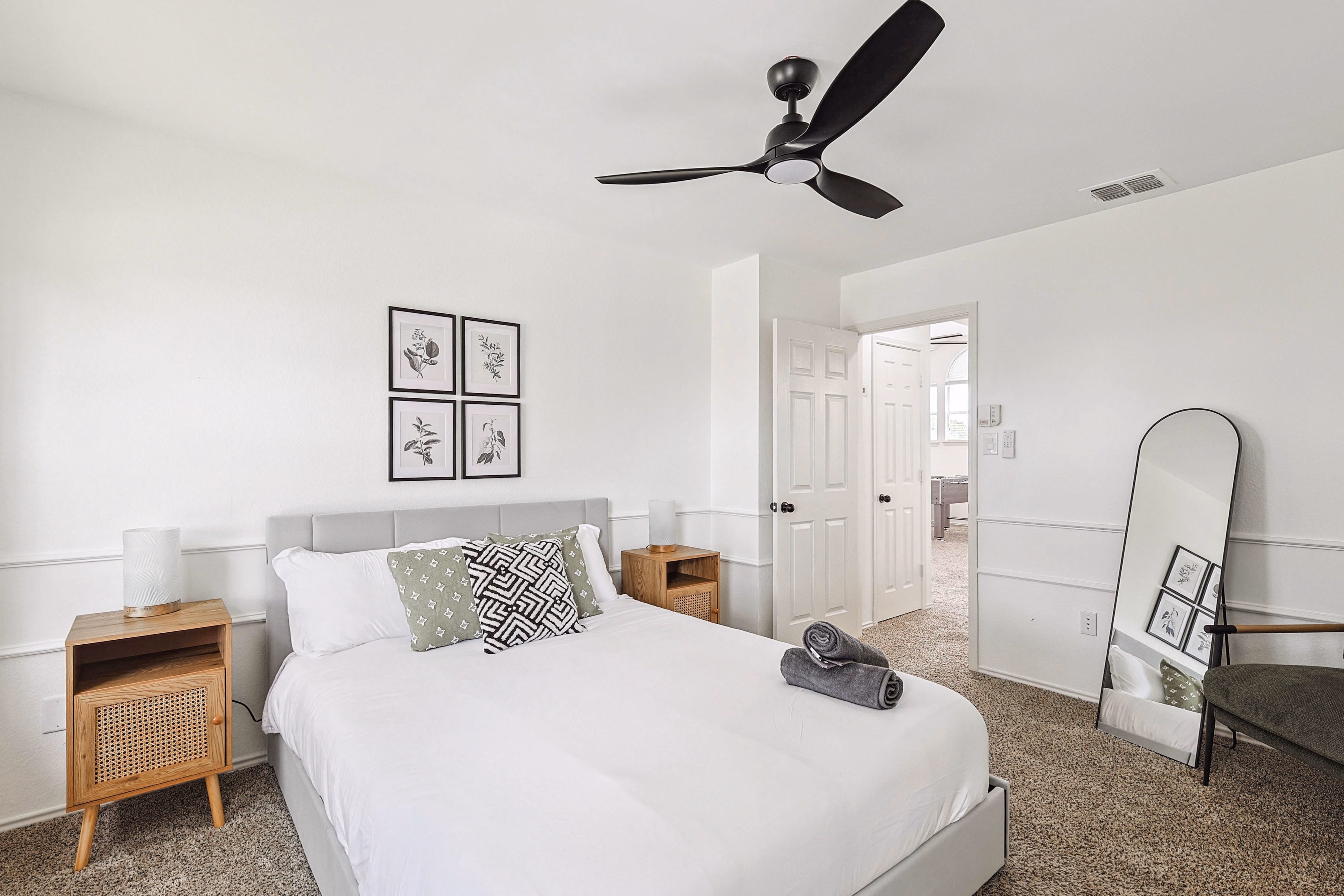 Upstairs bedroom — queen bed with 4-frame botanical art, twin rattan nightstands, view through doorway into hall.