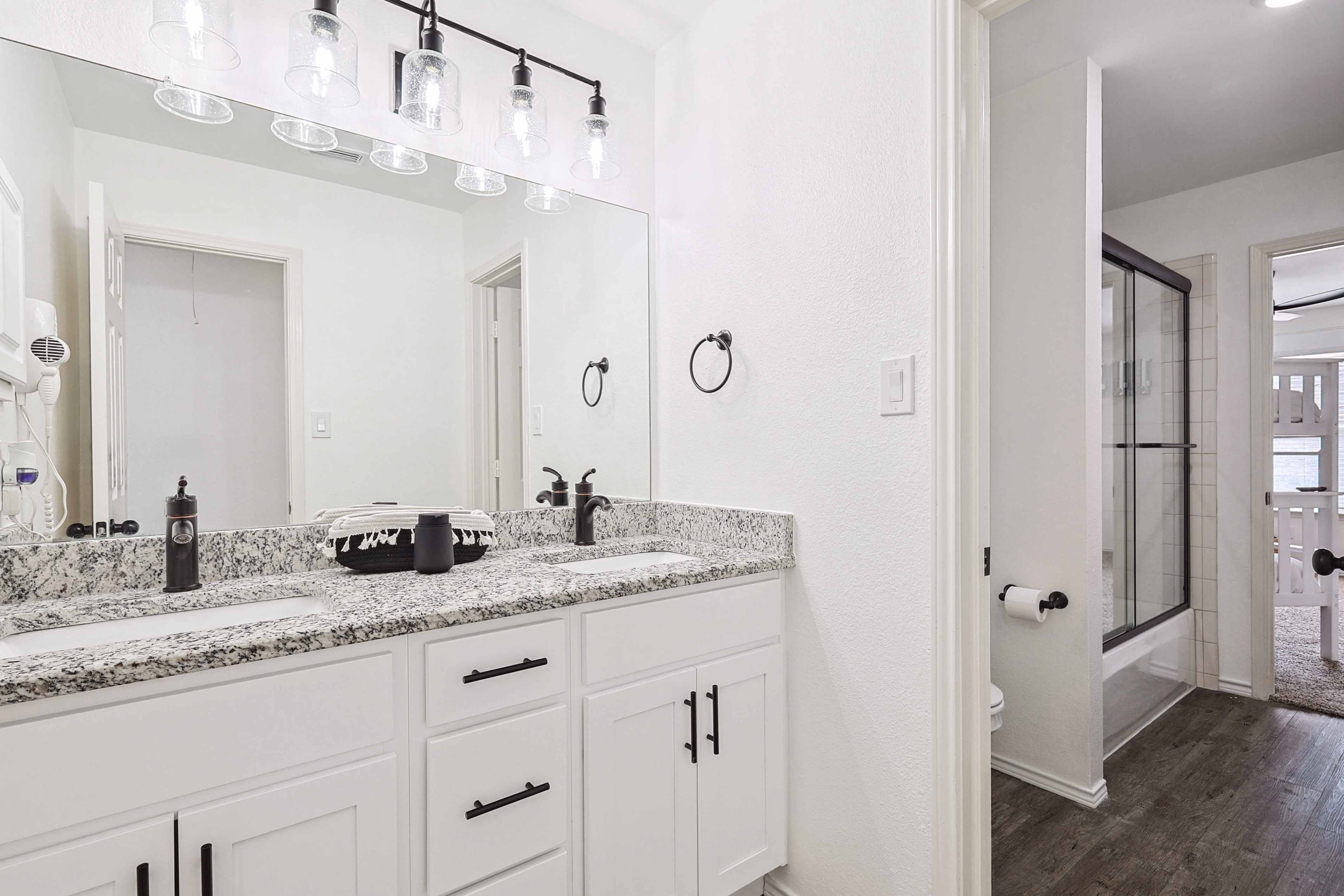 Upstairs bunk-room bathroom — double granite vanity with bronze fixtures, towel rings, sliding-door shower visible through doorway to bunk room.