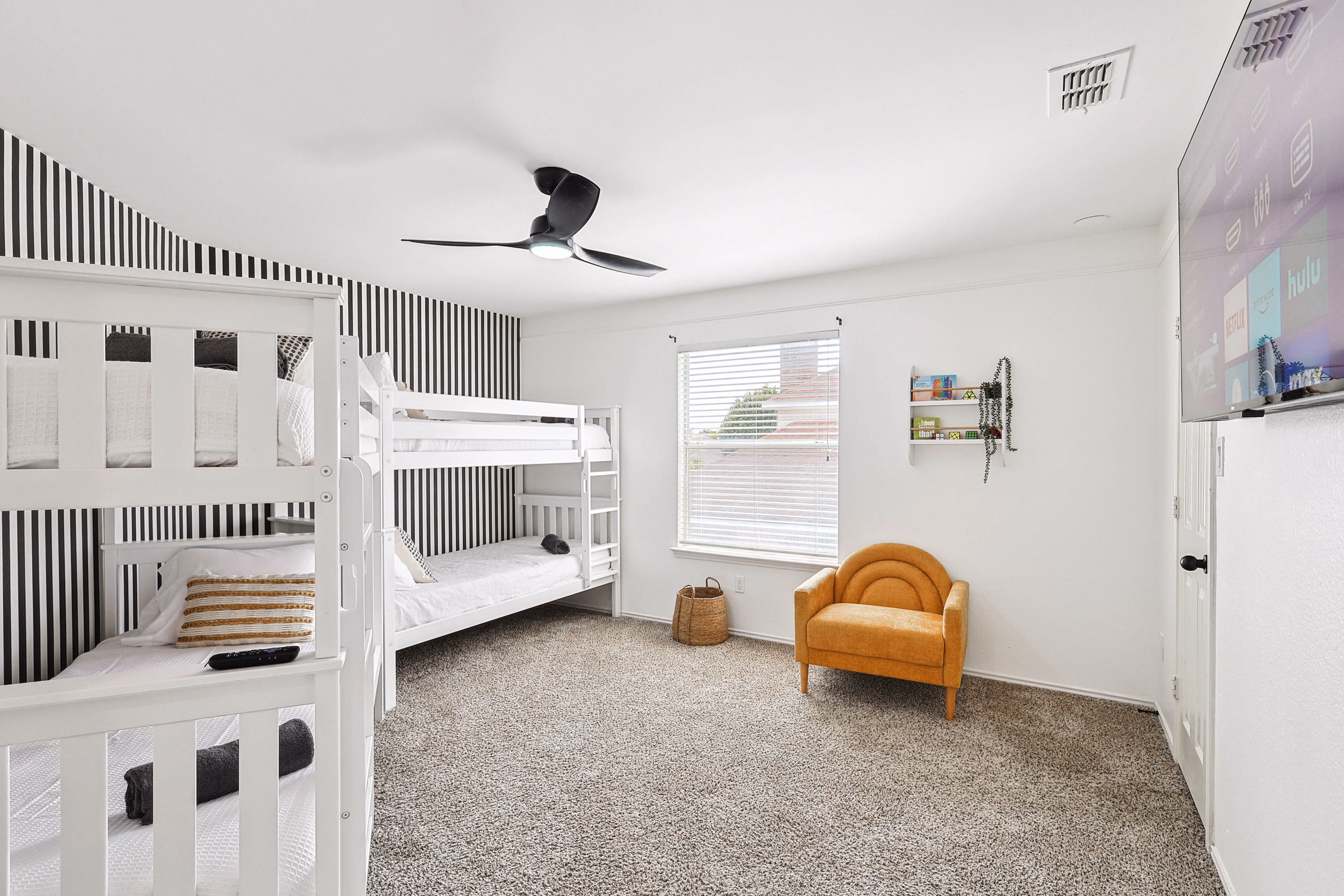 Bunk room — pair of stacked twin bunks against black-stripe wallpaper, orange accent chair, wall-mounted TV.