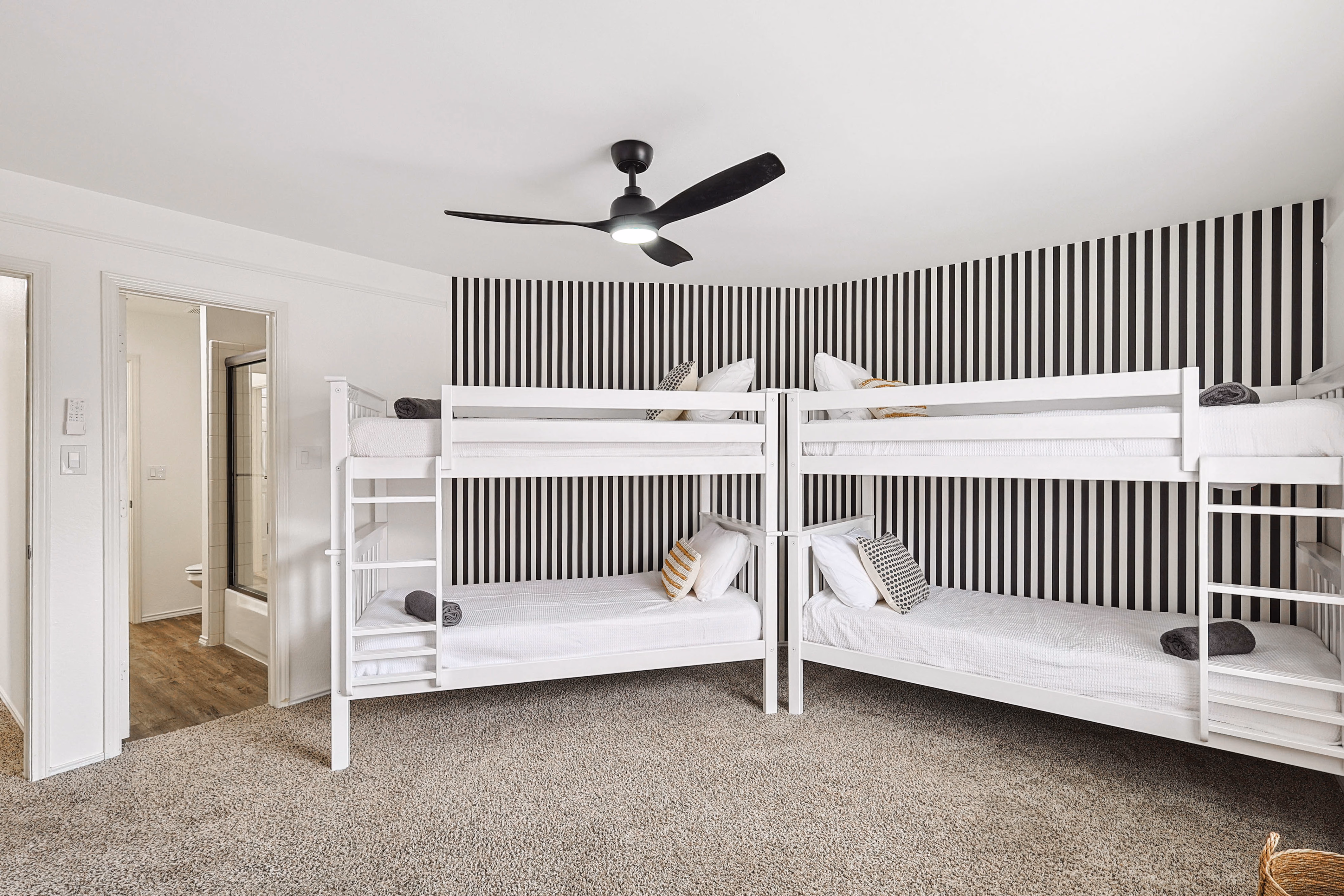Bunk room — four twin bunks in two side-by-side stacks, black-stripe wallpaper, ceiling fan, en-suite bath through left door.