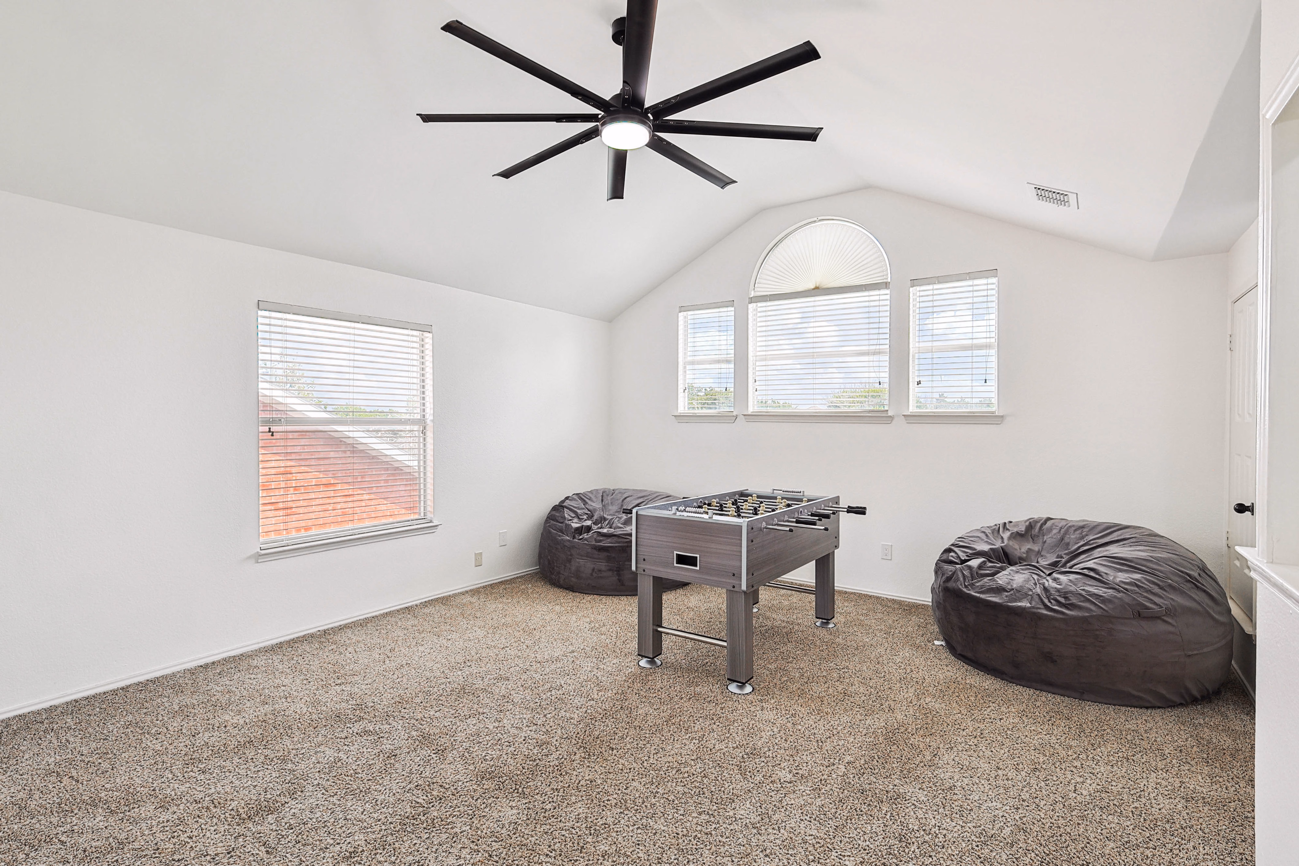 Loft game room — vaulted ceiling, oversized fan, foosball table flanked by oversized bean bags, half-moon arched window.