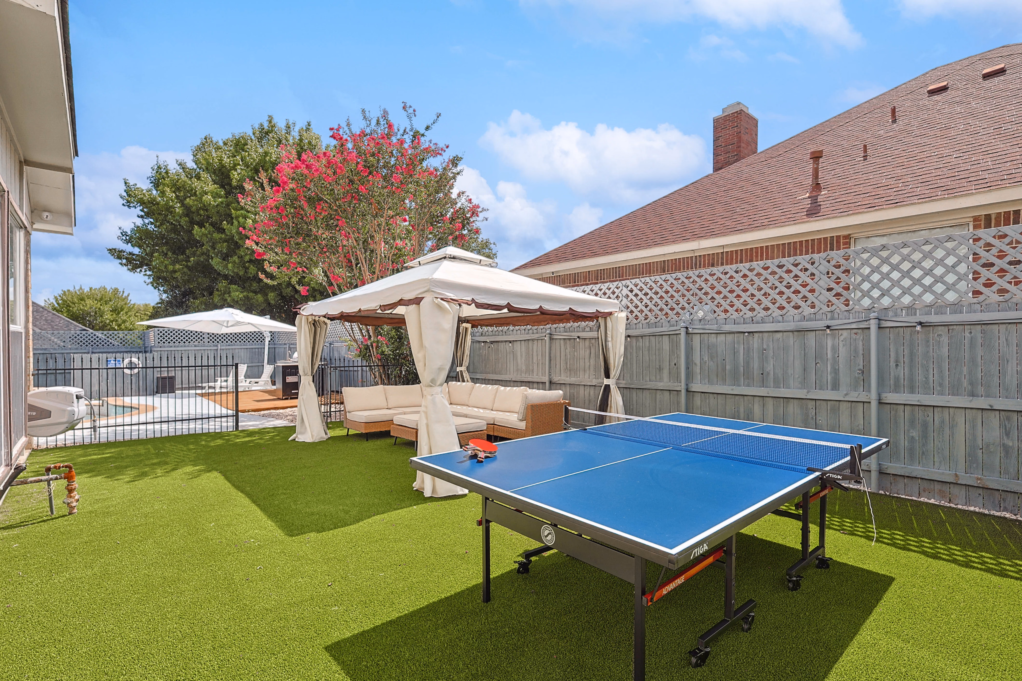 Side yard — full-size ping pong table on astroturf, canopy pavilion with sectional sofa beyond.
