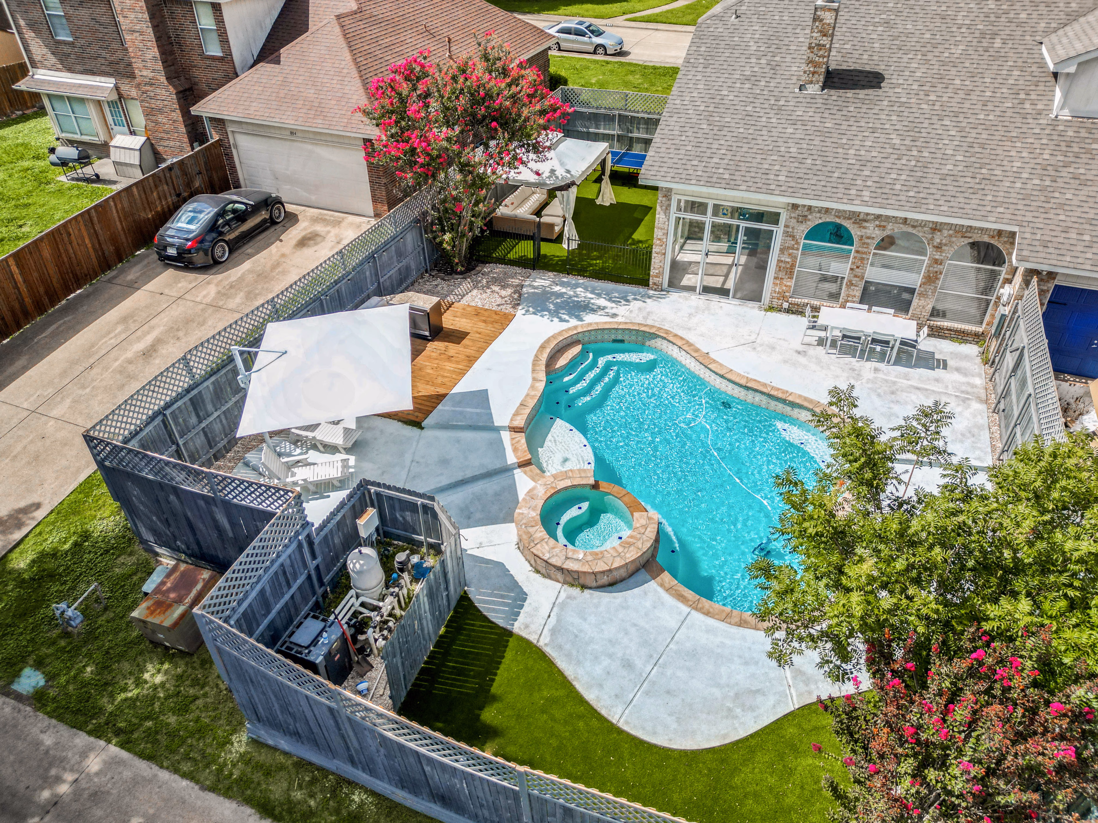 Aerial / drone — backyard from above showing pool with attached hot tub, outdoor kitchen, pavilion canopy and side yard.