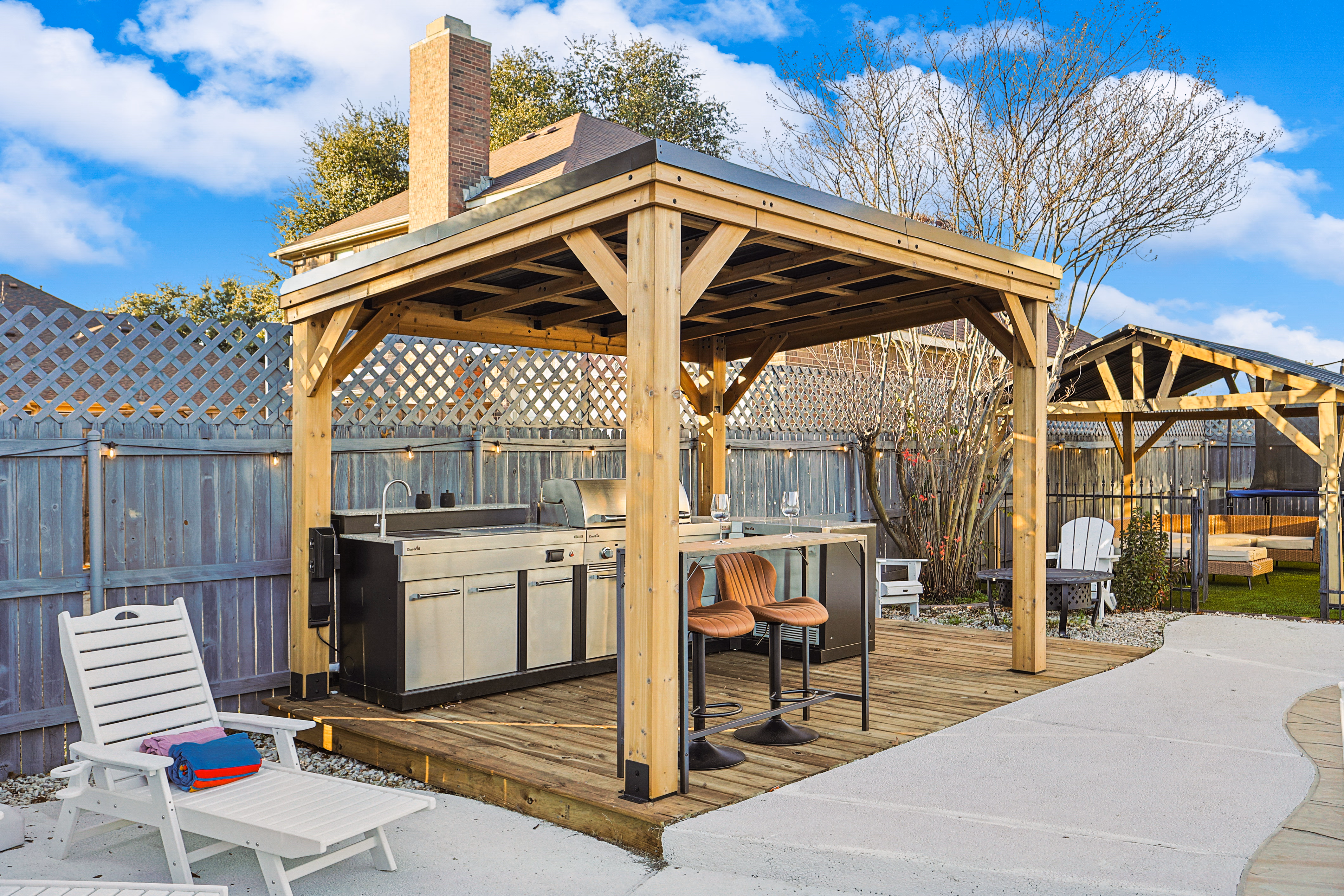 Wooden pavilion — built-in stainless outdoor kitchen with grill and gas griddle, lounge chair beside on the patio.