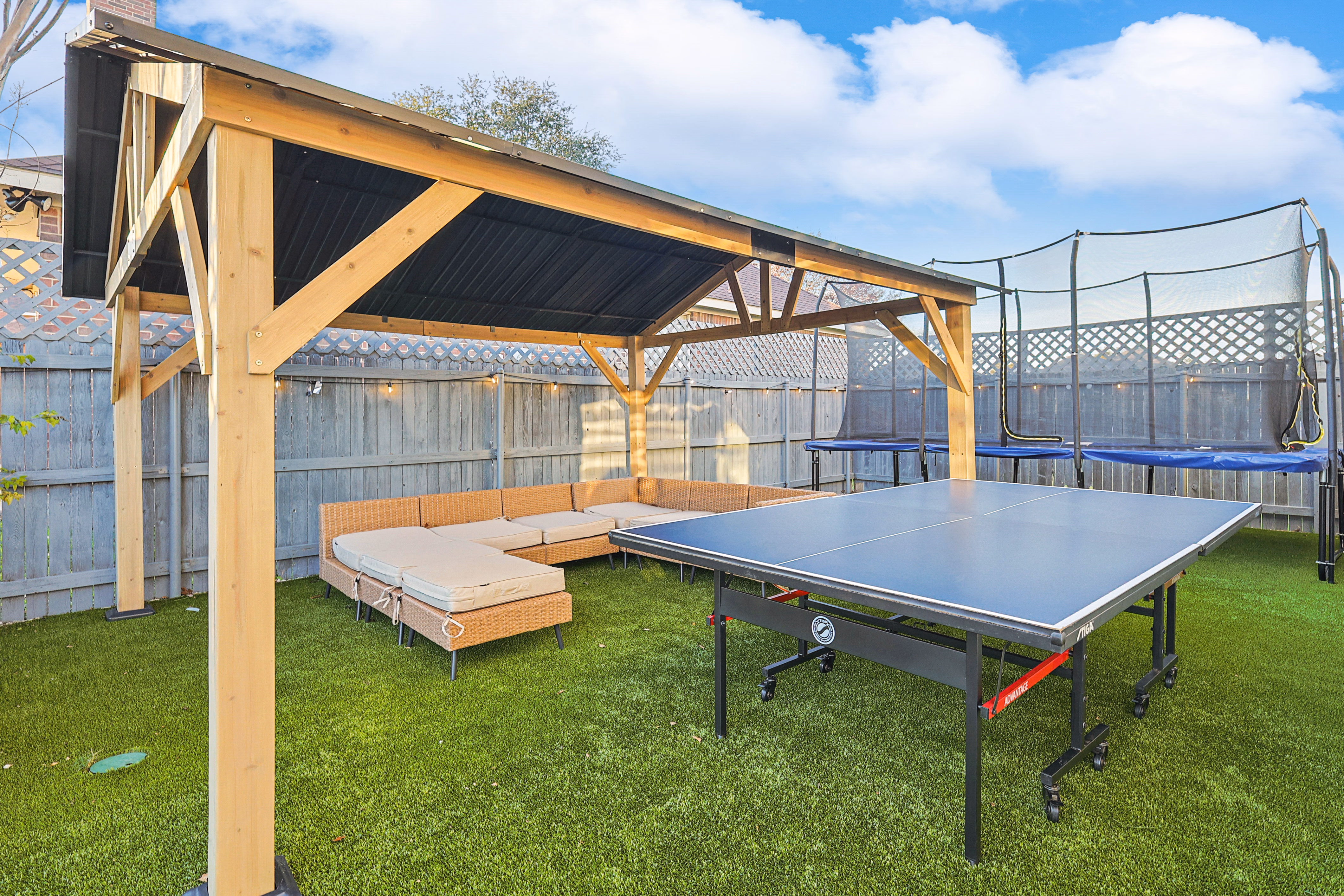 Wooden pavilion — sectional sofa under metal roof beside ping pong table on astroturf, full-size trampoline behind.
