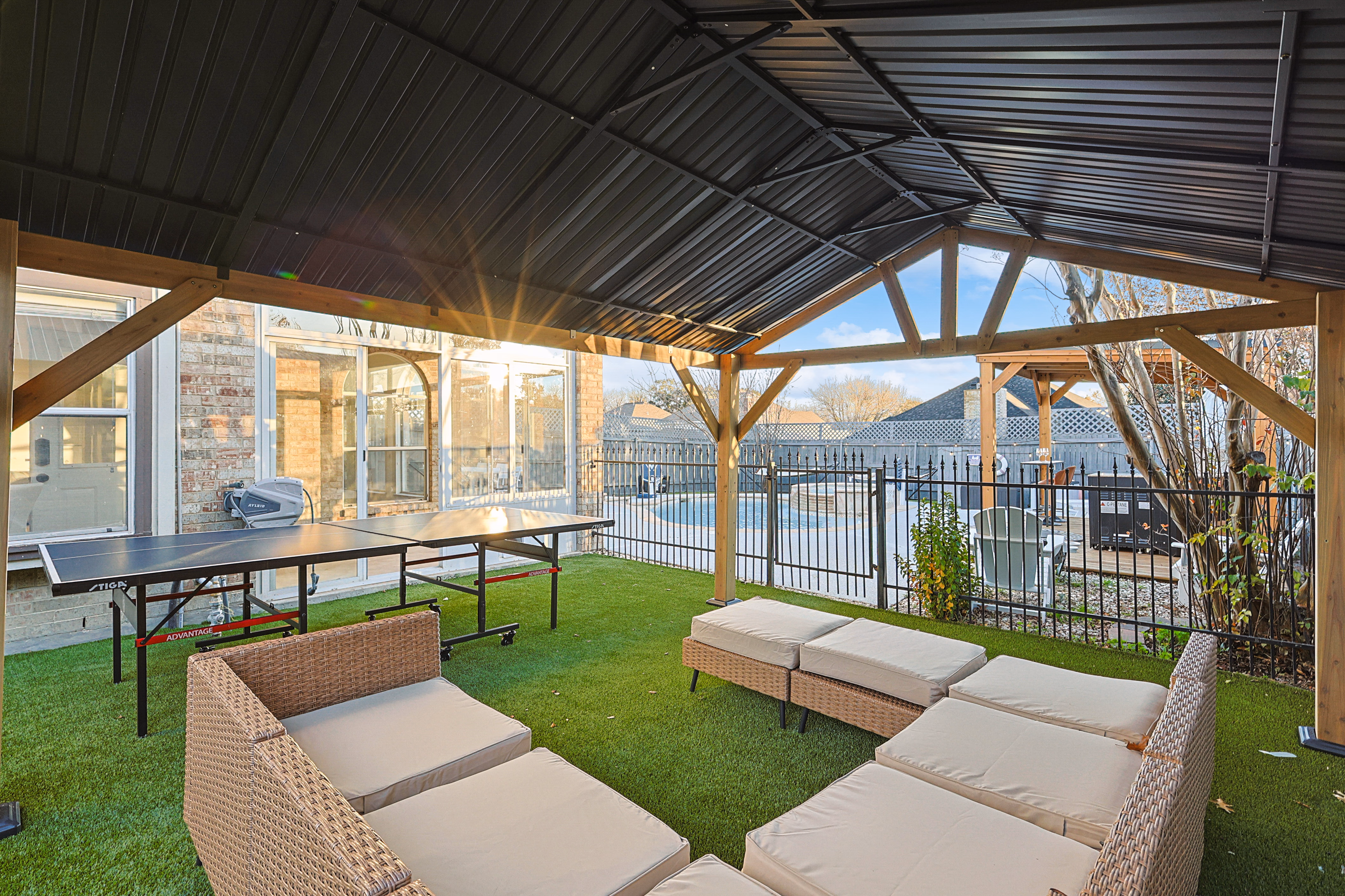 Wooden pavilion — ping pong table and sectional lounge looking out through the open roof posts toward the pool, sun streaming through.