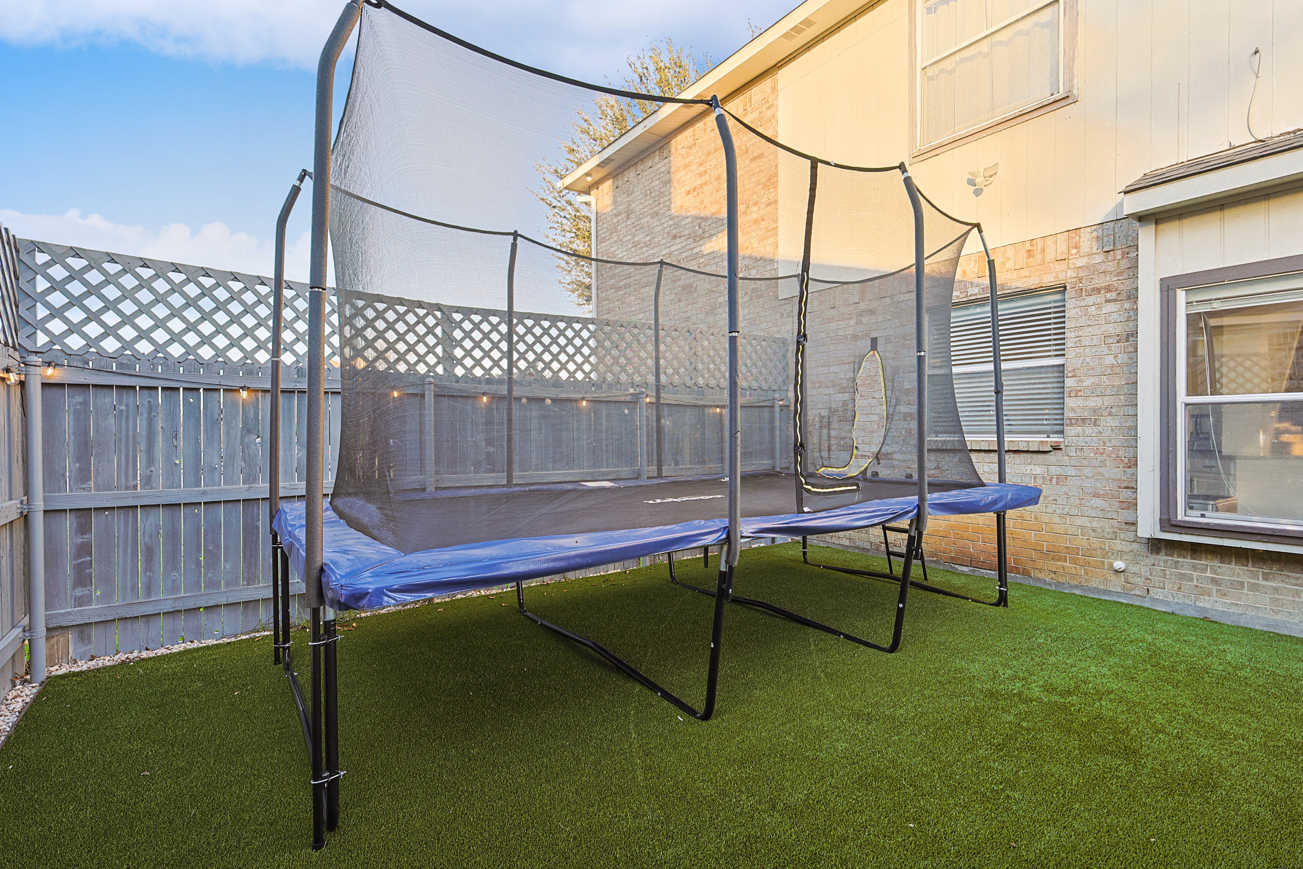Trampoline — full-size enclosed trampoline on astroturf side yard against lattice-topped fence.