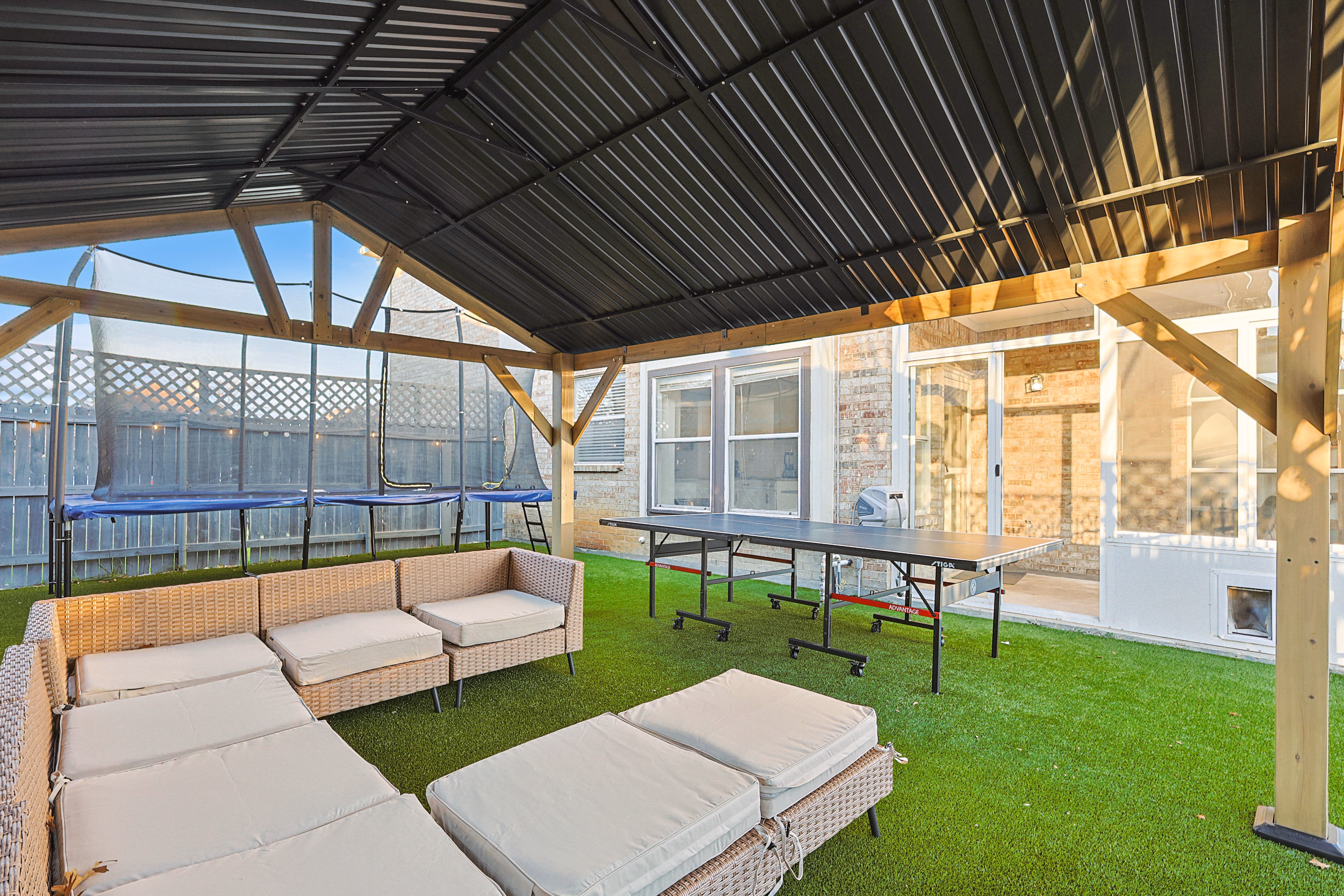 Wooden pavilion — lounge sectional, ping pong table and trampoline visible from the pool side.